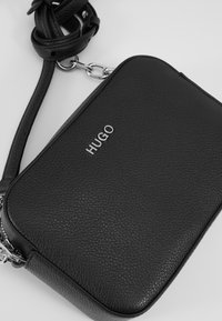 Černá kožená crossbody taška s texturovaným povrchem. Obsahuje stříbrné logo, nastavitelné ramenní popruhy a zipové uzavírání. Kompaktní obdélníkový design.