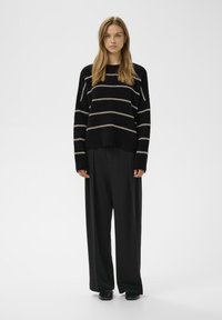 Suéter de rayas negras con rayas crema, de corte holgado y mangas largas, combinado con pantalones anchos negros y zapatos negros. Textura suave.