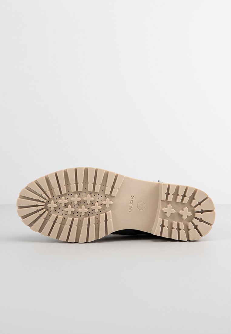 Schwarzes Schuhwerk mit einer strukturierten Gummisohle, die mit gemusterten Rillen versehen ist, und einer beigen Sohle. Logo auf der Sohle sichtbar.