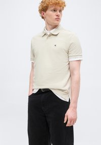 Jeune homme aux cheveux roux bouclés portant un polo beige Calvin Klein, un sous-maillot blanc et un pantalon noir, les mains dans les poches, regardant vers la droite.