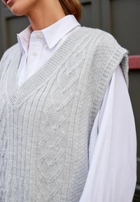 Gilet en tricot gris clair avec des motifs en point de câble, décolleté en V et bords côtes, superposé sur une chemise blanche à col.