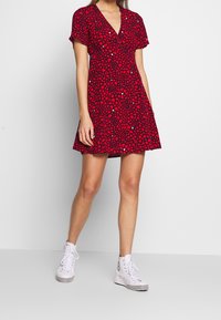 Vestido rojo con patrón de corazones en blanco y negro, mangas cortas y escote en V. Combinado con zapatillas altas blancas. Bajo corto y acampanado.