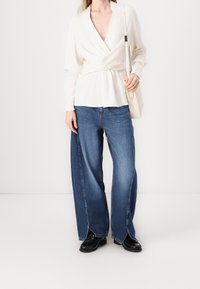 Biała bluzka z kopertowym dekoltem w kształcie litery V i długimi rękawami, w zestawieniu z szerokimi niebieskimi dżinsami z postrzępionym dołem oraz czarnym obuwiem.