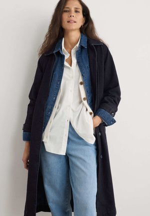 Langer schwarzer Mantel über einer Jeansjacke, weißem Hemd und hellblauen Jeans. Merkmale sind Knöpfe, ein auffälliger Kragen und gestaffelte Texturen.