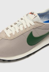 Sapatilha bege com materiais em malha e camurça. Apresenta um logo verde, atacadores brancos, colarinho almofadado e palmilha azul. Sola preta com ranhuras.