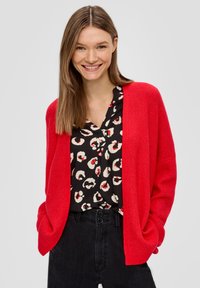 Rote gerippte Strickjacke mit offenem Vorderteil, getragen über einer schwarzen Bluse mit weißen und roten kreisförmigen Mustern, kombiniert mit schwarzen hoch geschnittenen Jeans.