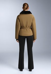 Brown padded jacket with a black fur collar, belted at the waist, paired with black flared pants featuring zip accents on the cuffs.