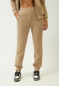 Beige Jogginghosen mit glatter Textur, elastischem Bund, seitlichen Taschen und schmal zulaufenden Beinen, kombiniert mit schwarz-weißen Turnschuhen.