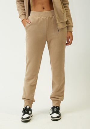 Pantalons de jogging beiges avec une texture lisse, taille élastique, poches latérales et jambes fuselées, associés à des baskets noires et blanches.