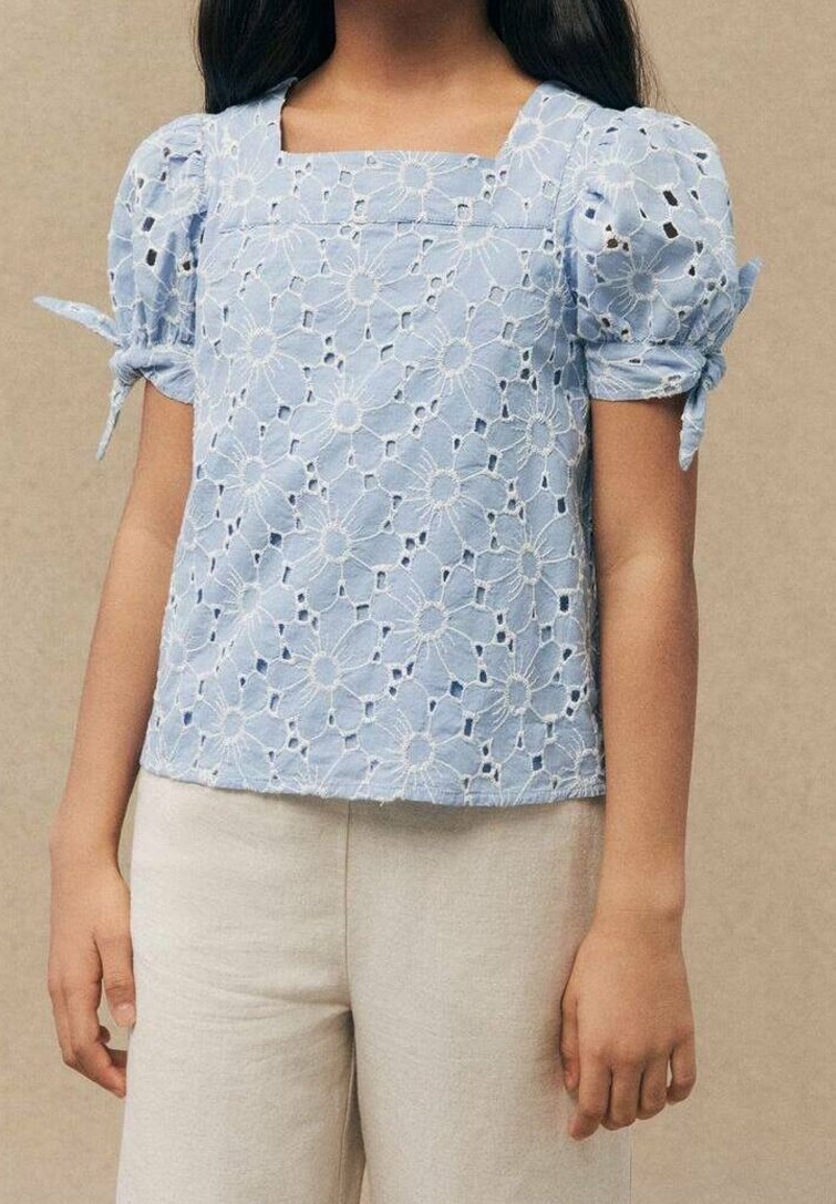 Fille portant un haut en dentelle ajourée florale bleu clair avec des manches bouffantes nouées aux coudes et un pantalon beige clair.