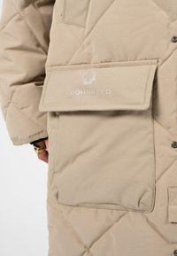 Béžová prošívaná bunda s velkým překrývacím kapsou vpředu označenou "PROHIBITED ENGINEERED BY STRELLSON", na které je vidět část ruky s prsteny.