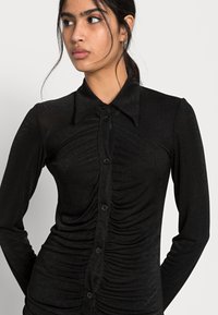 Camisa preta com botões, feita de tecido texturizado, apresentando um design ajustado com detalhes franzidos na parte da frente e um colarinho clássico.