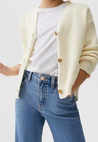 Person klädd i en vit strukturerad kofta med träknappar över en vanlig vit t-shirt och högmidjade blå jeans, med en hand i fickan.