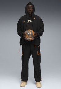 Sort hættetrøje med orange accenter, sort t-shirt og cargo-bukser med orange detaljer. Holder en mønstret basketball.