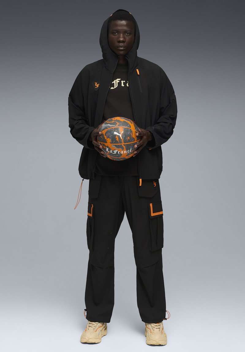 Mustast sportlikust komplektist koosnev kapuutsiga jakk oranžide detailidega, sobivad püksid koos cargo-taskutega ja beežid tossud, hoides korvpalli.