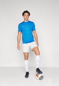 T-shirt de sport bleu à manches courtes avec des accents noirs, shorts blancs, chaussettes blanches avec un logo marine, et chaussures de football noires, se tenant à côté d'un ballon de football.