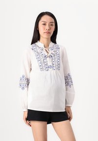 Blusa bianca con ricami blu, scollatura arricciata e maniche lunghe. Presenta una vestibilità comoda e dettagli decorativi sulle braccia.