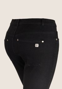 Vaqueros de mezclilla negra con un diseño ajustado, cintura de altura media, dos bolsillos traseros y detalles metálicos plateados. Textura suave y color oscuro.