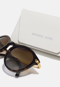 Aurinkolasit kilpikonnakuosisella rimalla, ruskeilla gradient-linsseillä, kultaisilla yksityiskohdilla ja tekstuurilla varustetulla valkoisella kotelolla, jossa on kohokuvioituna "MICHAEL KORS".
