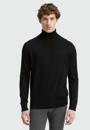 Maglione a collo alto nero con texture a coste, maniche lunghe e orlo aderente. Dettaglio della etichetta laterale che aggiunge un accento sottile. Indossato con pantaloni grigi.