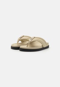 Beige flip-flop sandaler med en buet stroppdesign, med en metallisk detalj og en svart gummisåle for bedre grep og komfort.
