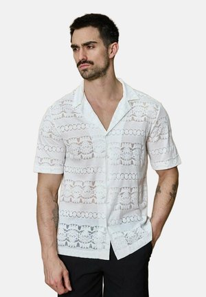 Homme portant une chemise blanche à manches courtes avec des motifs en dentelle et un pantalon noir, debout avec une main dans la poche.