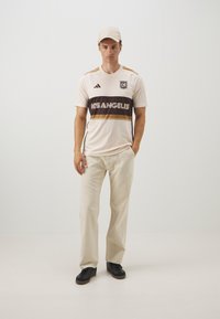 Maglietta sportiva crema con il testo "LOS ANGELES" in marrone e oro, dettagli neri, abbinata a pantaloni beige e sneakers nere, su uno sfondo neutro.