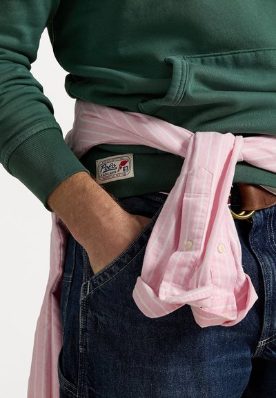 Felpa verde con polsini a coste, camicia a righe rosa annodata in vita e jeans scuri in denim con una cintura in pelle marrone.