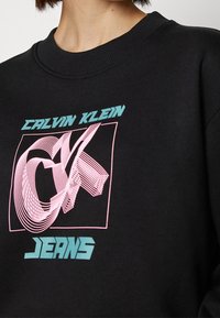 Černá mikina s grafickým designem, na které je nápis "CALVIN KLEIN" v tyrkysové barvě a růžový abstraktní vzor, označený "JEANS" také v tyrkysové.