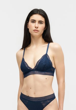 Kobieta o długich ciemnych włosach, ubrana w dopasowany granatowy koronkowy biustonosz i majtki na jasnym, jednolitym tle.