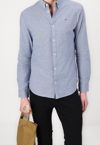 Camisa de botones azul claro con cuello, hecha de tela, con botones en la parte delantera y un logo en el pecho. Pantalones negros y un bolso de tela marrón.