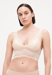 Lace bralette in nude, featuring a scalloped hem, wide lace straps, and a plunging neckline with floral patterns throughout.