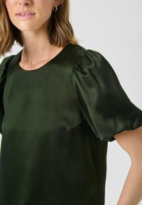 Blusa di raso verde scuro con maniche a palloncino corte, scollatura rotonda e una texture liscia. Vestibilità leggermente ampia, dettagli di design minimi.