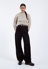 Vrouw met lange vlecht die een beige pluizige trui, brede zwarte broek, bruine riem en donkerbruine schoenen draagt, staand tegen een witte achtergrond.