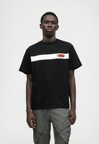 Camiseta de algodão preta com uma faixa horizontal branca e um patch de logo vermelho no peito. Mangas curtas, corte descontraído, textura suave.