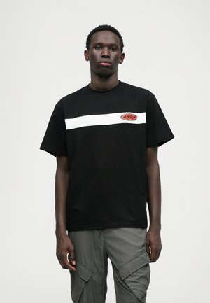 Czarny bawełniany t-shirt z białym poziomym paskiem i czerwoną łatą z logo na klatce piersiowej. Krótkie rękawy, luźny krój, gładka struktura.