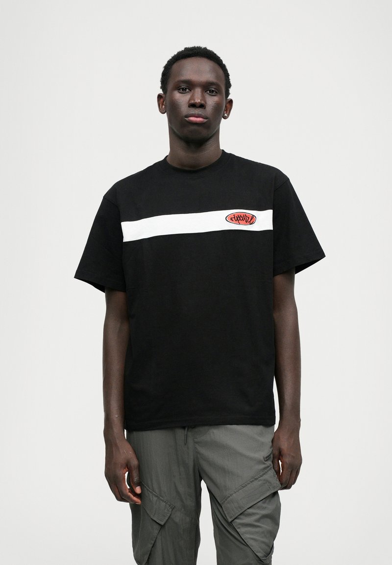 Camiseta de algodão preta com uma faixa horizontal branca e um patch de logo vermelho no peito. Mangas curtas, corte descontraído, textura suave.