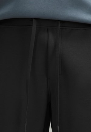 Schwarze Jogginghose mit glatter Textur, ausgestattet mit einem Kordelzugbund und dezenten Nahtdetails. Kombiniert mit einem blauen Oberteil oberhalb.