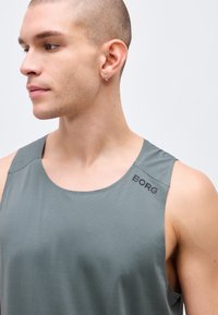 Νεαρός άνδρας με στενά ξυρισμένο κεφάλι που φοράει γκρι αμάνικο μπλουζάκι με το λογότυπο "BORG", κοιτάζει προς τα αριστερά σε απλό φόντο.