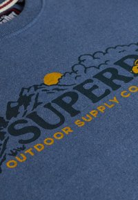 Blåt stof med teksten "Superdry Outdoor Supply Co", som har bjerg- og skymotiver med en gul sol delvist skjult bag bjergene.