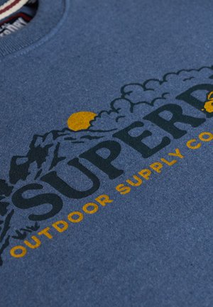 Tessuto blu con il testo "Superdry Outdoor Supply Co", caratterizzato da grafiche di montagne e nuvole con un sole giallo parzialmente nascosto dietro le montagne.