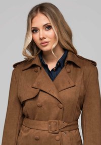 Abrigo trench doble abotonadura de ante sintético marrón con hombreras, cintura con cinturón y cuello con muesca, combinado con una camisa vaquera.