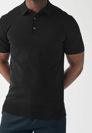 Mand iført en tætsiddende sort kortærmet polo med to knapper og mørkeblå bukser, står med hænderne bag ryggen.