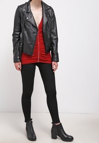 Kvinna i svart läderjacka, röd zip-up-tröja, svarta smala byxor och svarta klackade ankelstövlar som står mot en vit bakgrund.