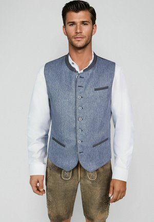 Homme portant une tenue bavaroise traditionnelle avec un gilet bleu à boutons, une chemise blanche et un short en cuir brodé marron, debout devant un fond uni.