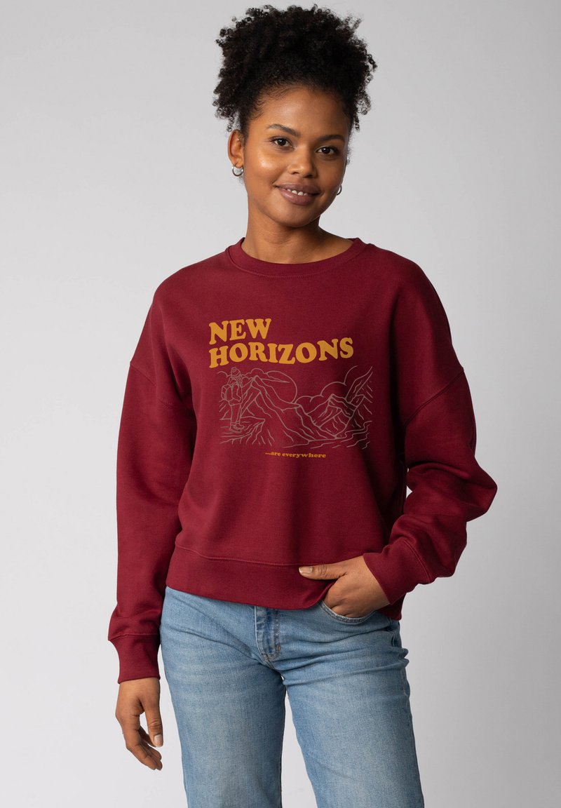 Bordeaux sweatshirt met "NEW HORIZONS" graphic in geel. Zachte stof, ontspannen pasvorm, geribde manchetten en tailleband, gecombineerd met lichtblauwe jeans.