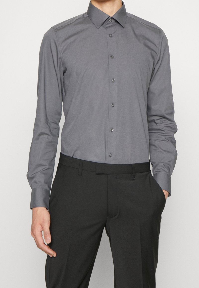 Homme portant une chemise grise ajustée à manches longues boutonnée, rentrée dans un pantalon de costume noir, debout avec une main dans la poche.
