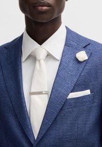 Blazer blu con texture, camicia bianca e cravatta. Presenta un fermacravatta argento, una spilla a forma di fiore bianca e un fazzoletto da taschino bianco.