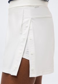 Λευκή φούστα με κουμπιά μπροστά και πλαϊνές στερεώσεις με αγκράφα που αποκαλύπτει λευκό σορτς από κάτω, φορεμένη σε σκουρόχρωμο πόδι.