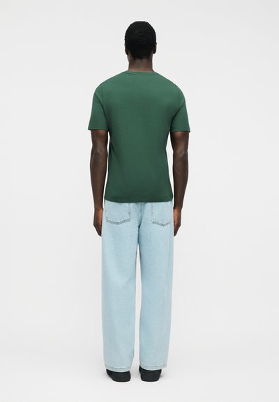 Άνδρας που φαίνεται από πίσω φοράει σκούρο πράσινο κοντομάνικο πουκάμισο, ανοιχτό μπλε φαρδιά τζιν παντελόνια και μαύρα παπούτσια με φόντο ένα απλό λευκό φόντο.